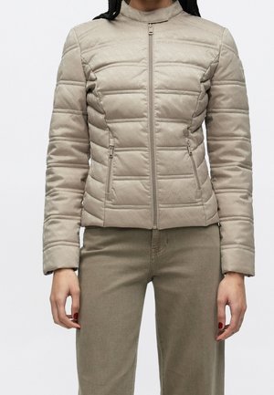 Kvinde iført en tætsiddende lys beige quiltet jakke med lynlåslommer og matchende beige bukser, stående mod en ensfarvet baggrund.