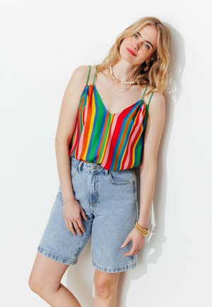 Femme blonde portant un haut sans manches rayé multicolore et un short en jean bleu clair, accessoirisée de colliers et de bracelets.