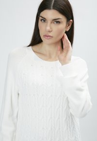 Weiße Strickjacke mit Kabelmuster, geripptem Halsausschnitt und Bündchen. Der Stoff wirkt weich und strukturiert, mit langen Ärmeln und einem lässigen Schnitt.