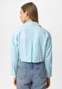 Lyseblå cropped skjorte med rygfald, lange ærmer og knapmanchetter. Bæres over højtaljede jeans med baglommer.