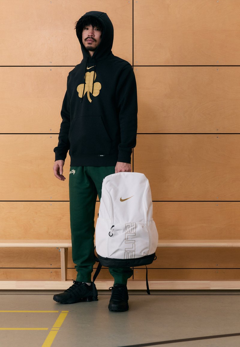 Bijeli Nike ruksak sa zlatnim detaljima i logotipom, elegantnog dizajna. Pamučna majica s kapuljačom s velikim zlatnim djetelinastim uzorkom, crna. Zelene sportske hlače.