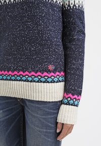 Marineblaues Strickpullover mit weißen, pinken und blauen gemusterten Säumen, gerippten Bündchen und Saum, getragen mit dunkelblauen Jeans.