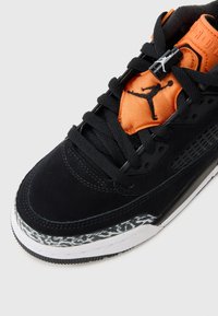Sneaker nera in suede con accenti arancioni, caratterizzata da un motivo testurizzato nella suola intermedia e da lacci prominenti. Logo visibile sulla linguetta.