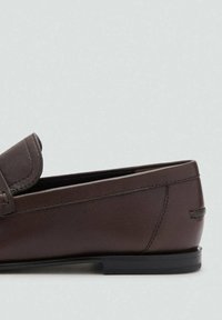 Mocasin de cuero marrón con punta redondeada, costuras decorativas y suela plana negra. La parte superior presenta una solapa decorativa.