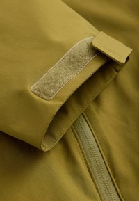 Material de culoare verde măsliniu, închidere cu velcro și un fermoar neted. Textură moale cu un finisaj mat, tăiat aproape, pentru a evidenția detaliile de design.