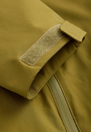 Tejido en color verde oliva, cierre de velcro y una cremallera suave. Textura suave con un acabado mate, cortado de cerca para enfatizar los detalles del diseño.