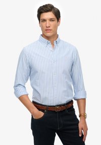 Lysblå stribet skjorte med knapper, lange ærmer, krave og brystlogo; båret med mørke jeans og et flettet brunt bælte.