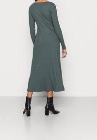 Kvinna i en långärmad, ribbad, mörkgrön midiklänning som har svarta klackstövlar, stående mot en enkel vit bakgrund.