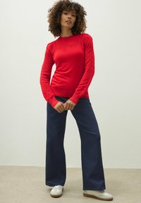 Kobieta z kręconymi włosami, ubrana w strukturalny czerwony sweter, granatowe spodnie o szerokich nogawkach i białe sneakersy, stojąca na tle gładkiej ściany.