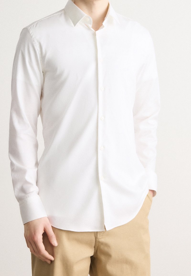 Homme portant une chemise blanche unie à manches longues boutonnée avec un pantalon beige, une main dans la poche, debout devant un fond blanc.