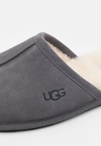 Szürke velúr papucs krémszínű birkabőr béléssel és gumitalppal. Az oldalon UGG logó található. Egyszerű dizájn.