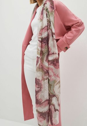 Femme portant un long manteau rose, un haut blanc, un pantalon blanc et une grande écharpe à motifs floraux dans des tons roses, blancs et verts.
