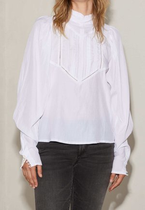 Vrouw draagt een witte blouse met lange mouwen en kantdetails en zwarte jeans, staand tegen een effen lichte achtergrond.