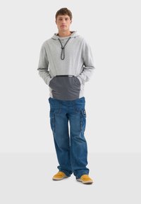 Sudadera gris con un bolsillo gris, capucha con cordón y pantalones cargo azules informales. El modelo lleva zapatillas amarillas. Diseño simple con elementos funcionales.