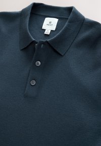 Dark navy polo shirt made of soft fabric, featuring a three-button placket, flat collar, and a tag labeled "NEXT" under the collar.
