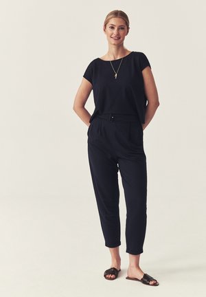 Kobieta stojąca z rękami za plecami, ubrana czarny krótki top, spodnie z wysokim stanem i paskiem, czarne sandały typu slide oraz złoty naszyjnik z zawieszką.