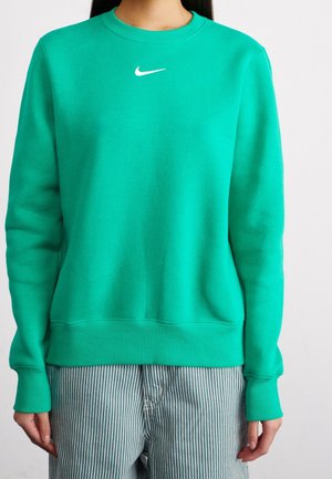 Femme portant un sweat-shirt Nike vert vif avec un logo blanc, associé à un pantalon à rayures verticales bleu et blanc.