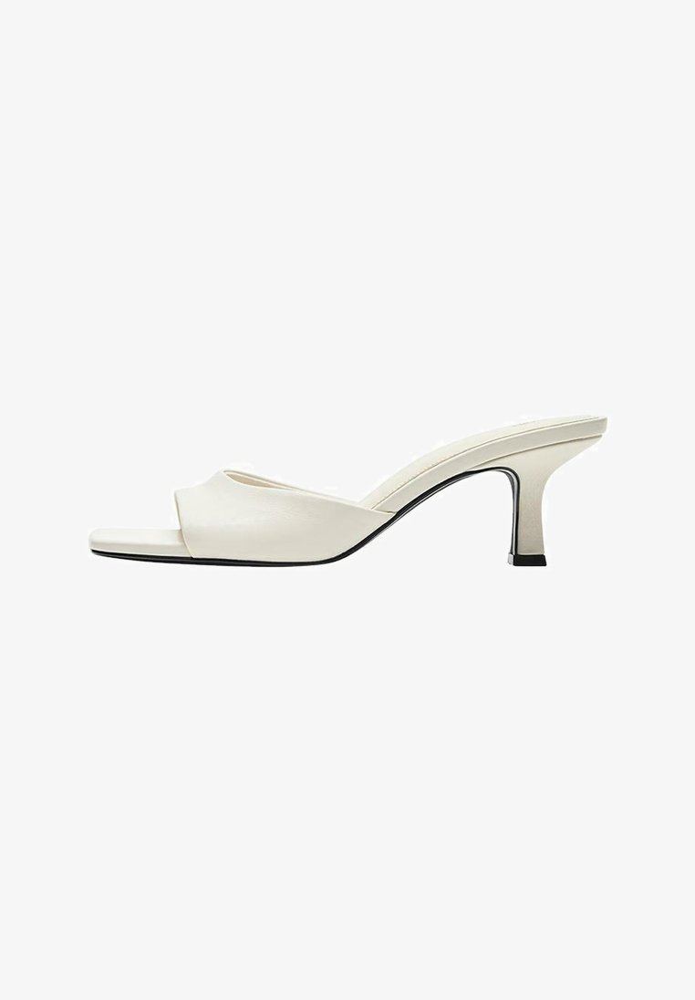Mule en cuir blanc à bout ouvert avec un talon bloc incurvé de hauteur moyenne et une semelle carrée légèrement effilée à l’avant.