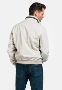 Lichtbeige bomberjack met een staande kraag, geribbelde manchetten en zoom, met contrasterende strepen. Draagt over blauwe jeans.