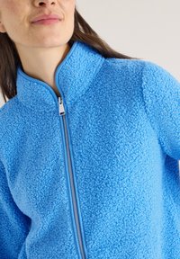 Femme portant une veste polaire bleu vif texturée avec une fermeture éclair frontale, sur un fond clair uni.