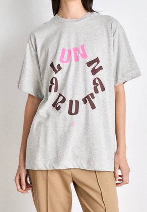 Person iført en grå t-shirt med teksten "UN NATURAL" i cirkulære lyserøde og brune bogstaver, kombineret med beige bukser.