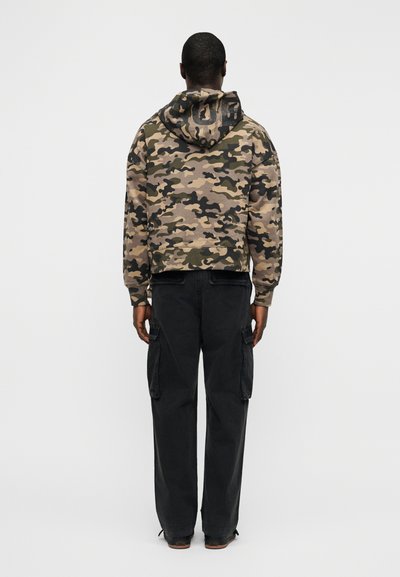 Φούτερ με κουκούλα σε καμουφλάζ χρώματα μπεζ, πράσινο και καφέ, συνδυασμένο με μαύρο cargo παντελόνι με πλαϊνές τσέπες, σε θέα από πίσω.