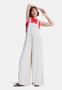 Salopette en lin couleur crème avec jambes larges, bretelles ajustables et poches avant, portée sur un haut rouge sans manches. Des sandales marron complètent la tenue.