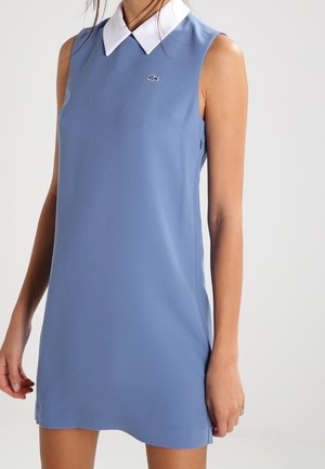 Sleeveless blue dress with white pointed collar, small embroidered logo on chest, worn by a standing person against a plain background.