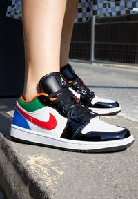 Sneakers de perfil bajo con parte superior de cuero sintético blanco, acentos negros, superposiciones en rojo y verde, y una lengüeta en azul. Suela negra texturizada.