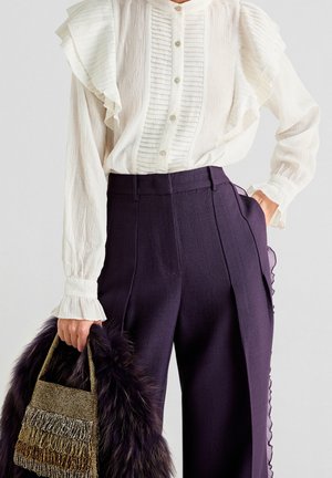 Frau trägt eine weiße Rüschenbluse mit langen Ärmeln und hoch taillierte dunkelviolette Hose, hält eine schwarze und goldene Fransenhandtasche.