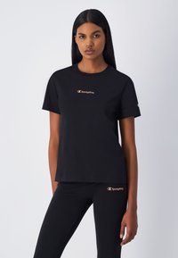 T-shirt di cotone nero con collo rotondo, caratterizzata da un logo Champion arancione sul petto. Ambientata su uno sfondo semplice.