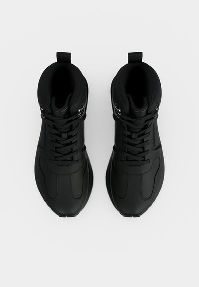 Ζευγάρι μαύρα sneakers ψηλά, εμφανισμένα από πάνω σε ανοιχτό φόντο, με κλείσιμο με κορδόνια και επενδεδυμένο κολάρο.