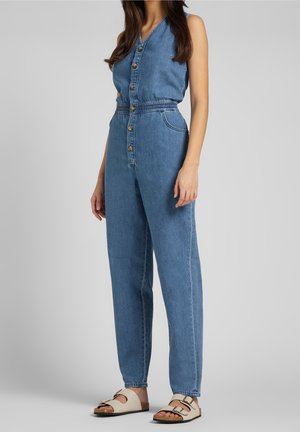 Kvinde iført blå denim-ærmeløs overall med frontknapper og beige bøjlesandaler, stående mod en ensfarvet baggrund.