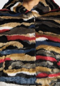 Manteau en fausse fourrure multicolore avec des rayures horizontales en noir, beige, blanc, rouge et bleu, porté par une personne visible du cou vers le bas.