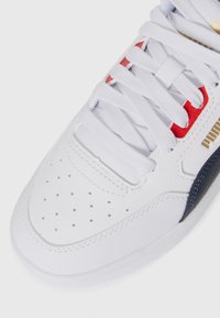 Sneaker in pelle bianca con accenti rossi e blu navy, caratterizzato da dettagli perforati sulla punta, lacci piatti e un logo dorato sul lato.