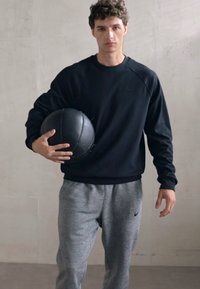 Sweatshirt de sport noir avec des manchettes côtelées, un pantalon de survêtement gris et un ballon de médecine noir, le tout sur fond de mur minimaliste.