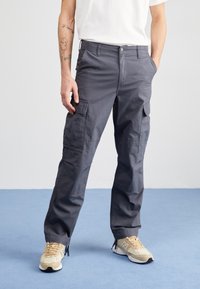 Dunkelgraue Cargo-Hosen mit lockerer Passform, mehreren Taschen und sichtbaren Nähdetails. Kombiniert mit beigen Sportschuhen.