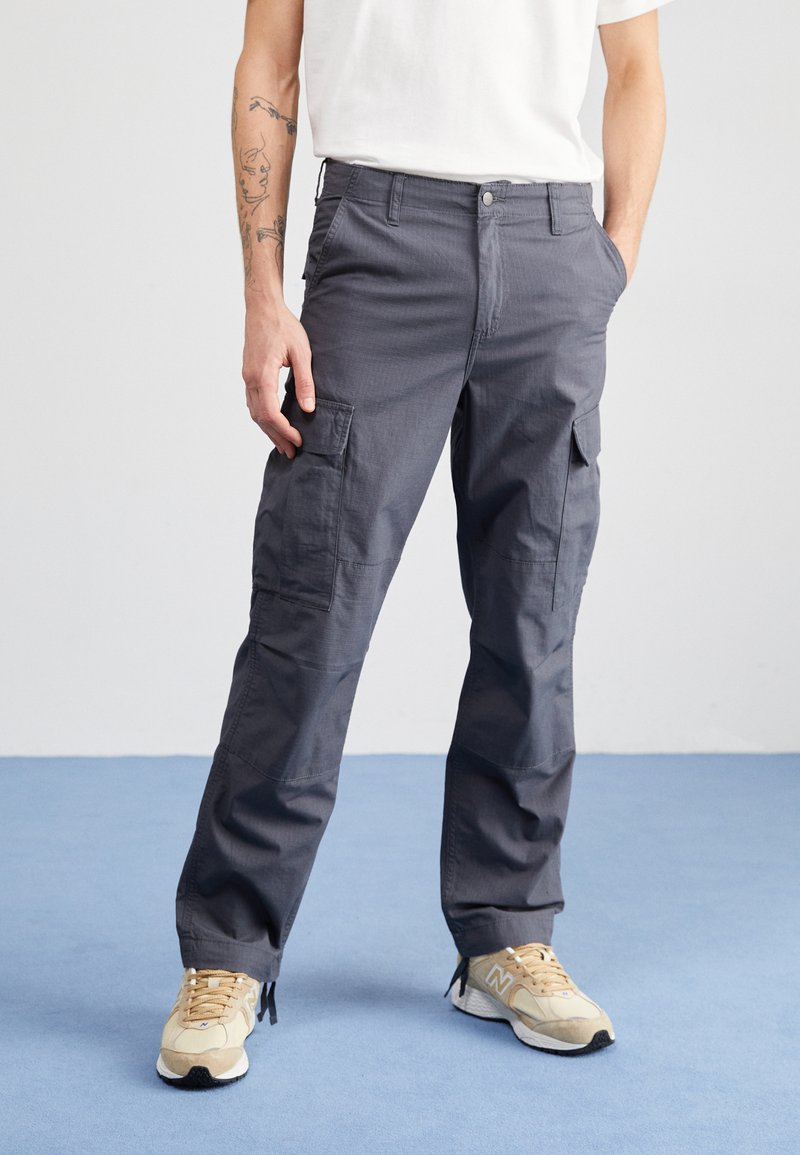 Dunkelgraue Cargo-Hosen mit lockerer Passform, mehreren Taschen und sichtbaren Nähdetails. Kombiniert mit beigen Sportschuhen.
