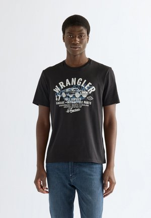Giovane uomo in piedi davanti a uno sfondo bianco, indossa una maglietta nera Wrangler con grafica e jeans blu, guarda avanti con un'espressione neutra.