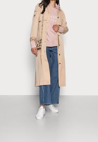 Gabardina beige con detalles de botones sobre una sudadera con capucha rosa claro, combinada con vaqueros de pierna ancha azul y zapatillas blancas. Bolso con estampado animal como toque de acento.