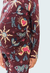 Top de manga larga color marrón oscuro y pantalones con un patrón colorido de soles, pájaros, libélulas, corazones y acentos florales. Textura de tela suave.