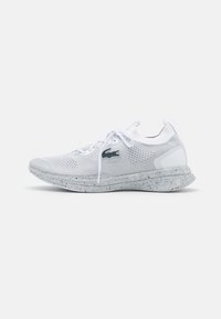 Chaussure de sport blanche en mesh avec des accents gris, tige texturée, lacets étroits, et semelle tachetée. Logo Lacoste sur le côté extérieur.