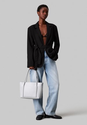 Femme portant un blazer noir ceinturé sur un bralette noir, jean large bleu clair, chaussures noires, tenant un grand sac fourre-tout blanc, debout sur un fond gris.