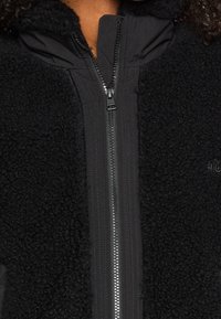 Zbliżenie na czarną kurtkę polarową z centralnym zamkiem błyskawicznym oraz teksturowanym materiałem przypominającym wełnę na przednim panelu i kołnierzu.