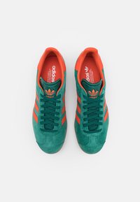 Zapatillas de ante verde con acentos naranjas y tres rayas. Presentan una superficie texturizada, puntera redonda y cierre con cordones.