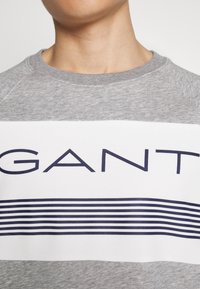 Persona che indossa un felpa grigia e bianca con il logo "GANT" blu navy e strisce orizzontali sul petto.