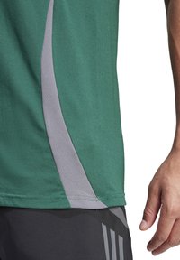 Maglietta sportiva verde con pannello laterale grigio. Il tessuto è texturizzato e presenta un design con collo a giro. Una mano è parzialmente visibile, posata in vita.