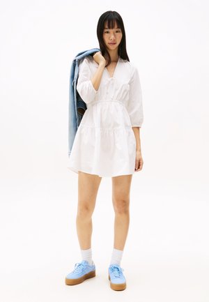 Vestito bianco a maniche corte con vita arricciata e balze, abbinato a sneakers azzurro chiaro e giacca di jeans appoggiata sulla spalla.