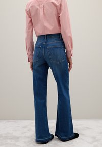 Persona che indossa jeans a vita alta a zampa d'elefante blu e una camicia a maniche lunghe a righe rosse e bianche infilata dentro, in piedi su un tappeto chiaro.