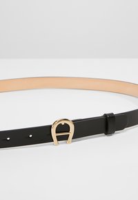 Cinturón de cuero negro con una hebilla en tono dorado con forma de bocado, que presenta una textura suave y múltiples orificios de ajuste a lo largo de su longitud.